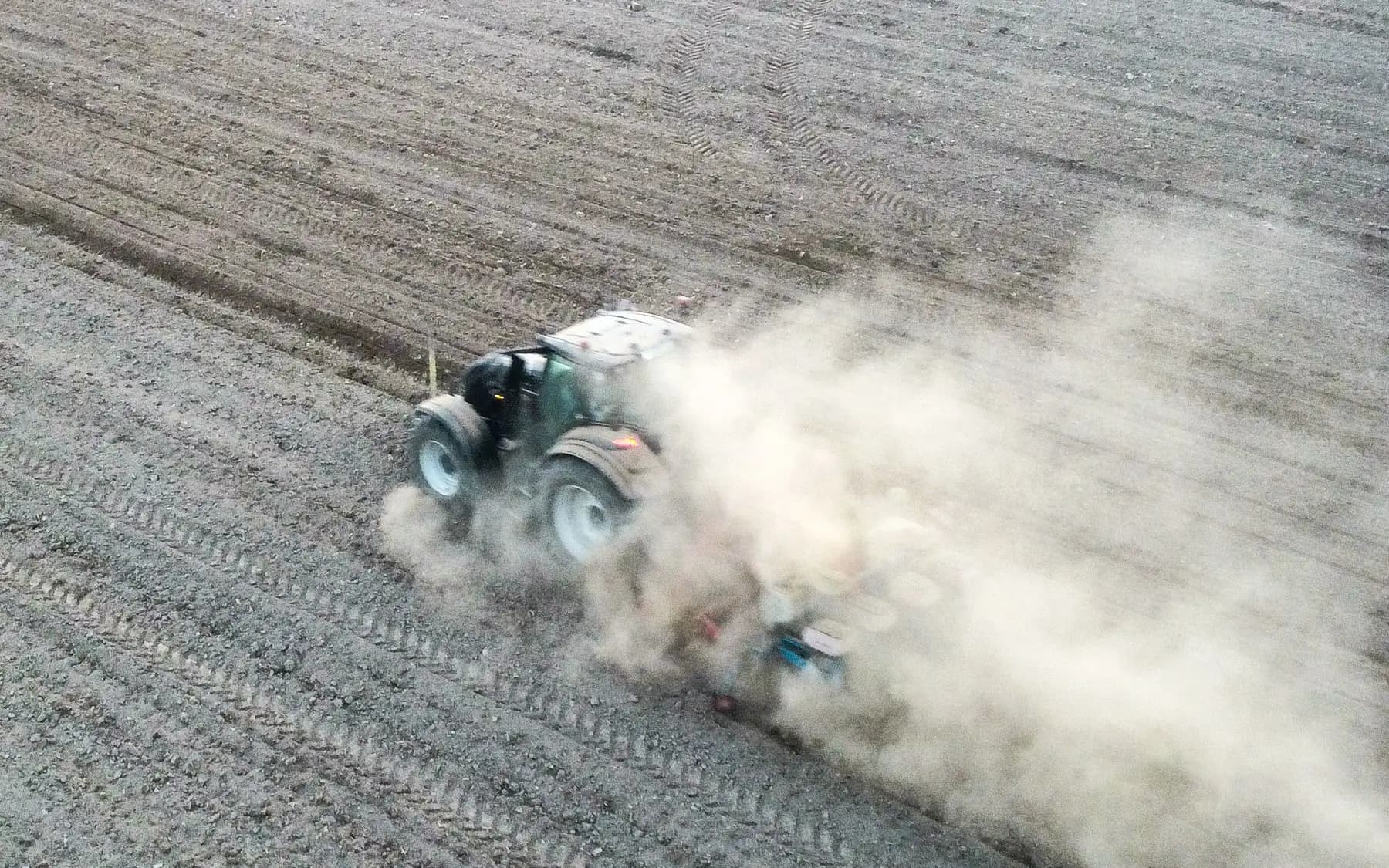 Traktor som drar ett redskap över ett torrt fält, sett uppifrån, med ett dammoln bakom sig.